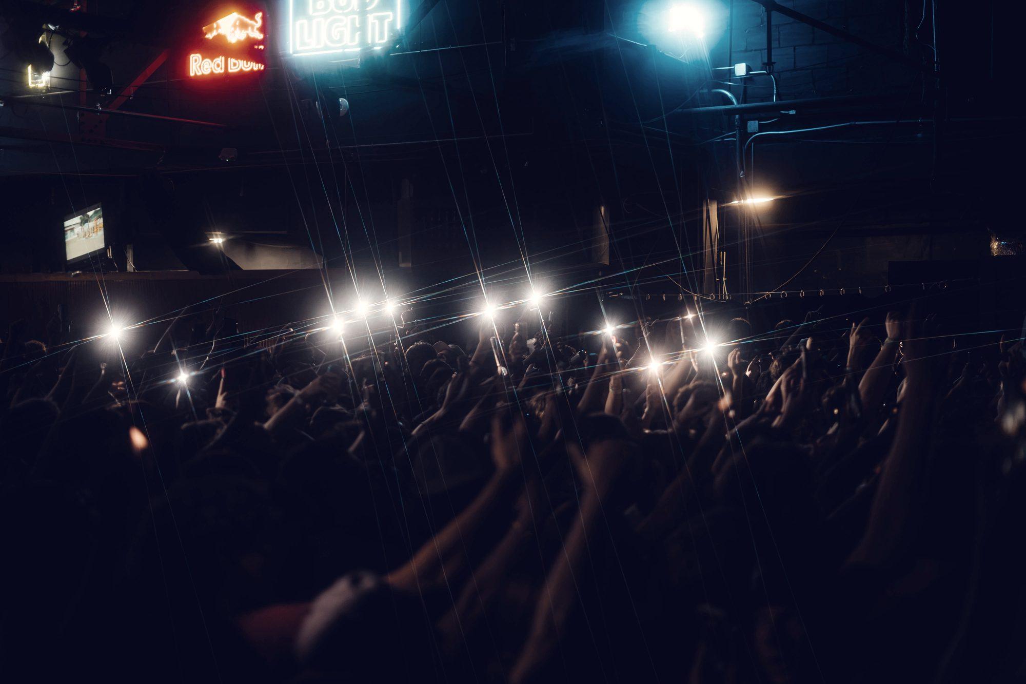 The crowd photographed from the back of the room, phone lights up and neon signs on the wall above.