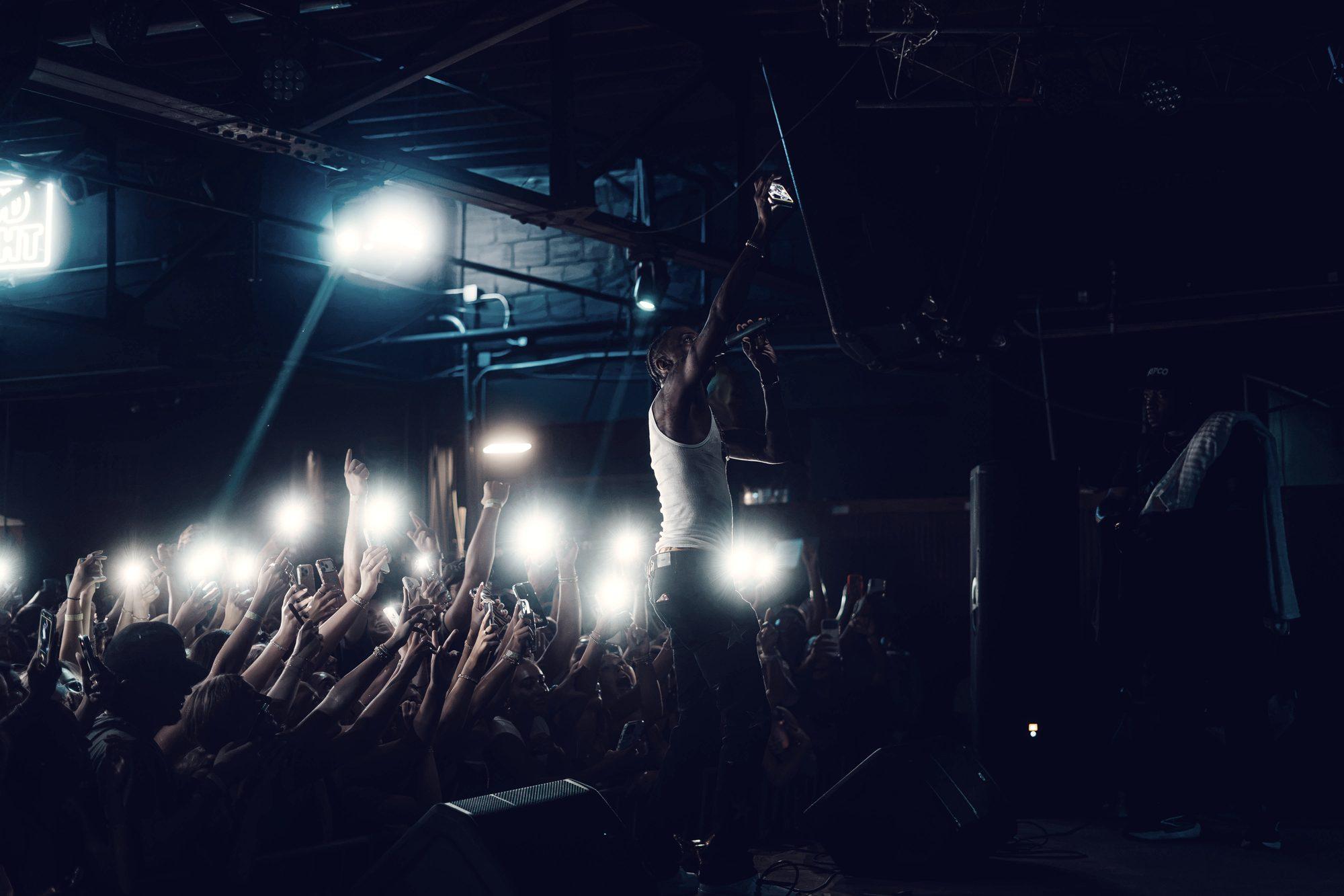 A performer at the front of the stage with one arm raised, lit from behind while the front rows hold their phones overhead.